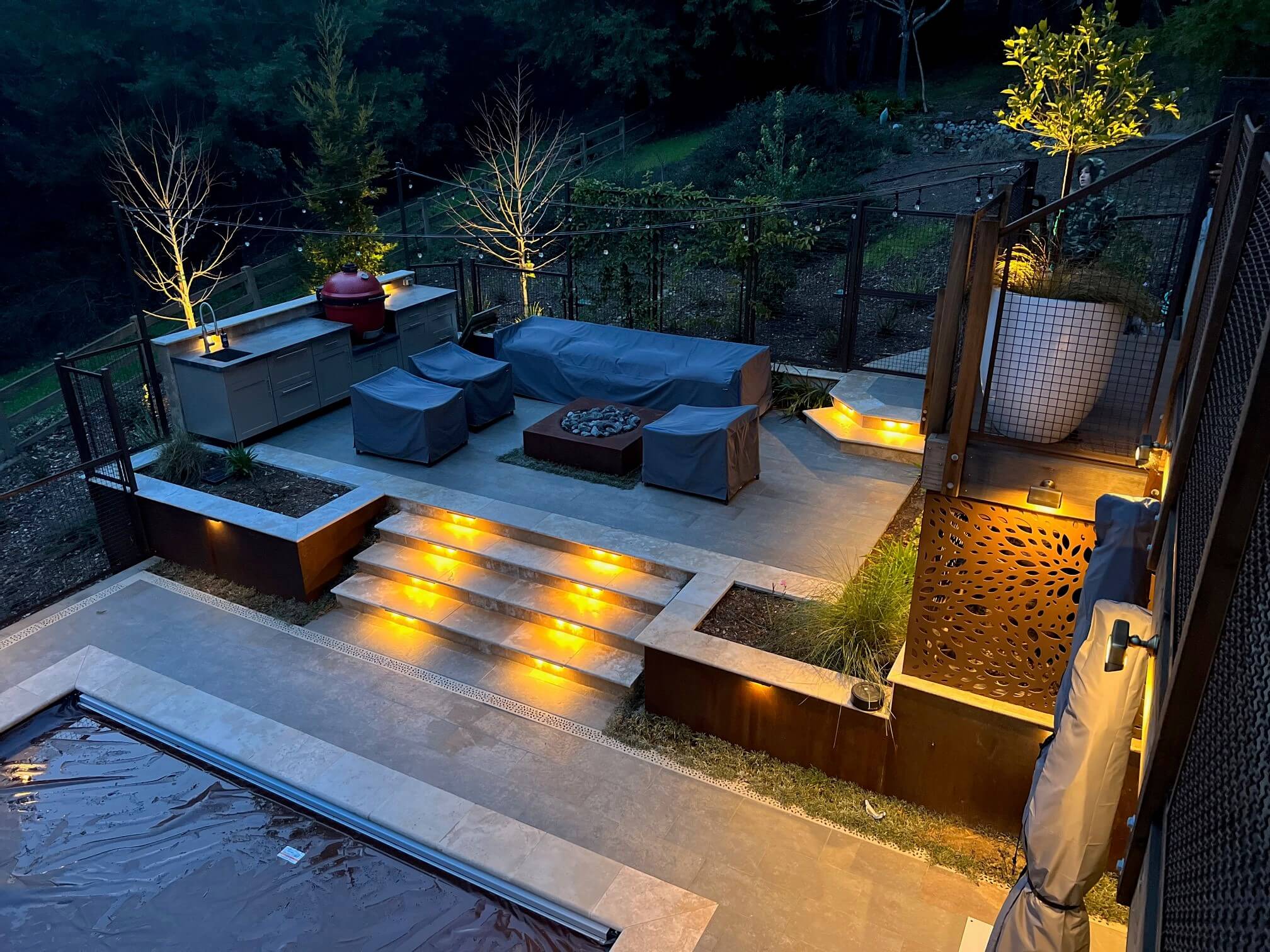 Nightime view of a firepit seating area on a raised patio terrace of a sloped backyard