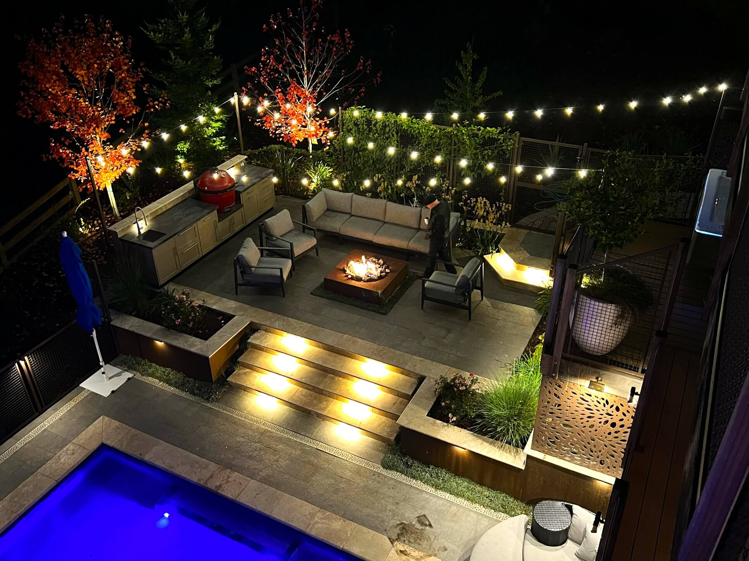 Outdoor kitchen and firepit area at night on steep sloped backyard with swimming pool