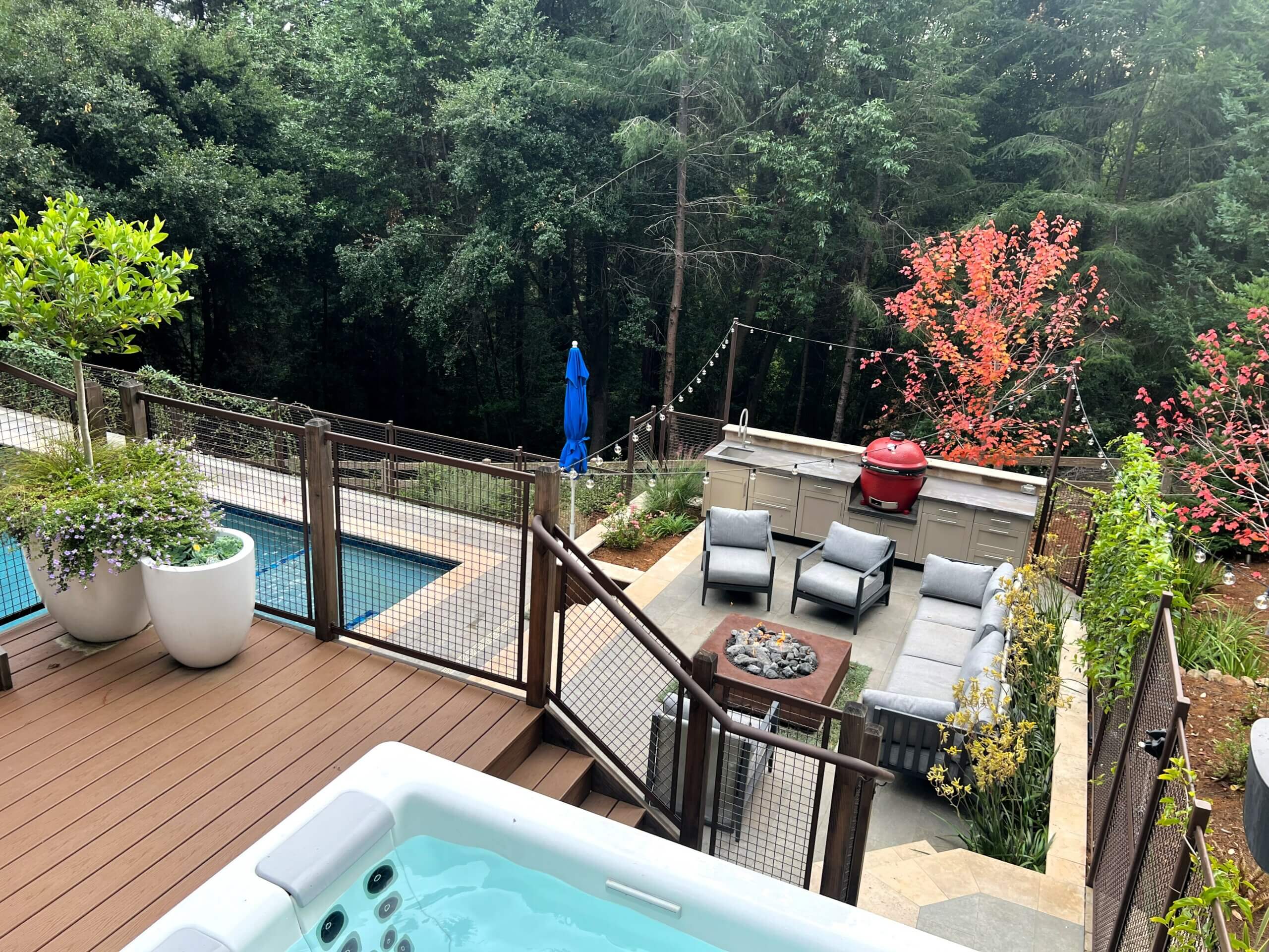 View of seating area from upper deck showing firepit, pool and hillside forest in background