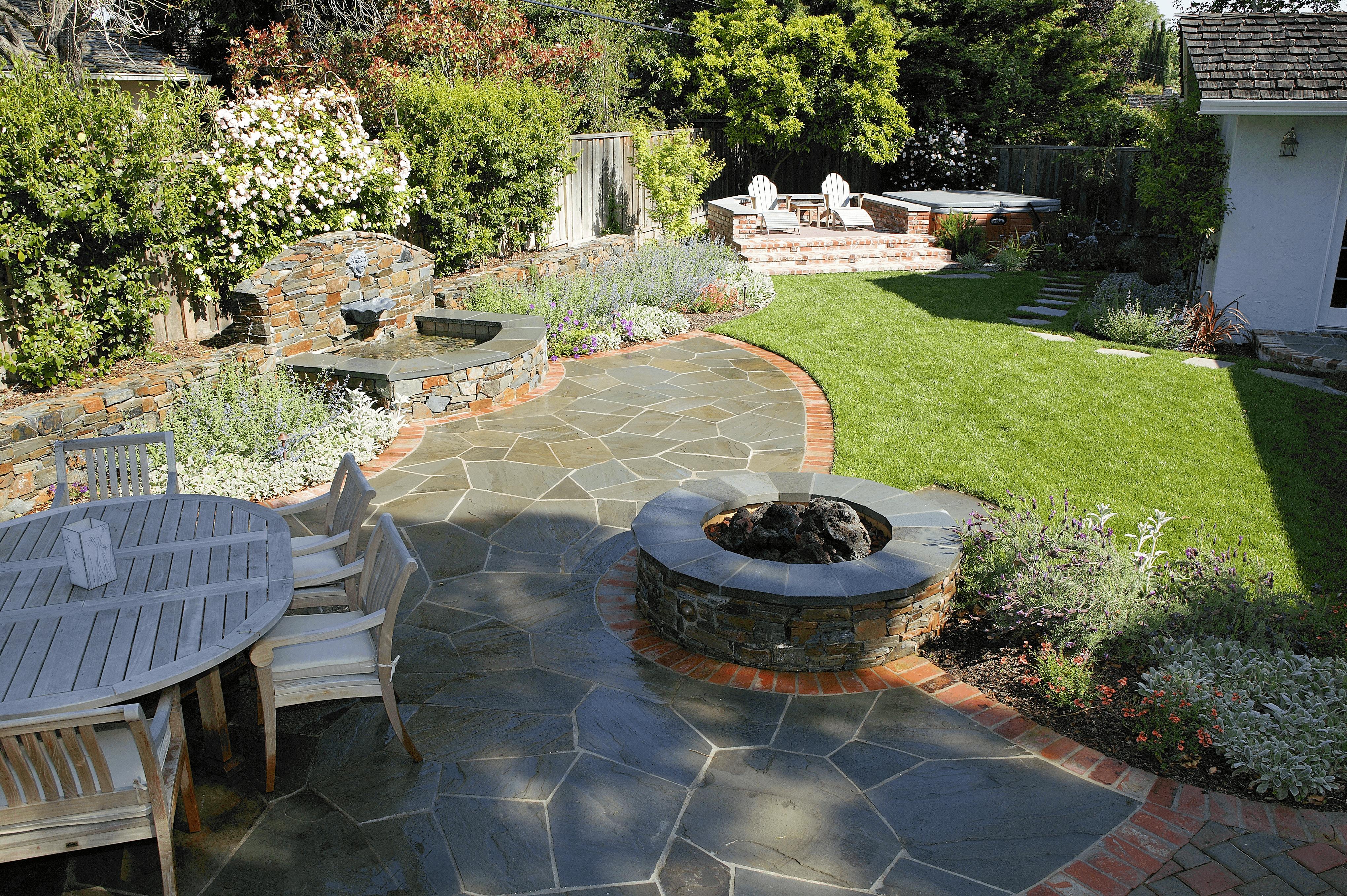 Firepit, seating area, and water feature amongst pavers and a lush lawn in a cottage-style California backyard landscape