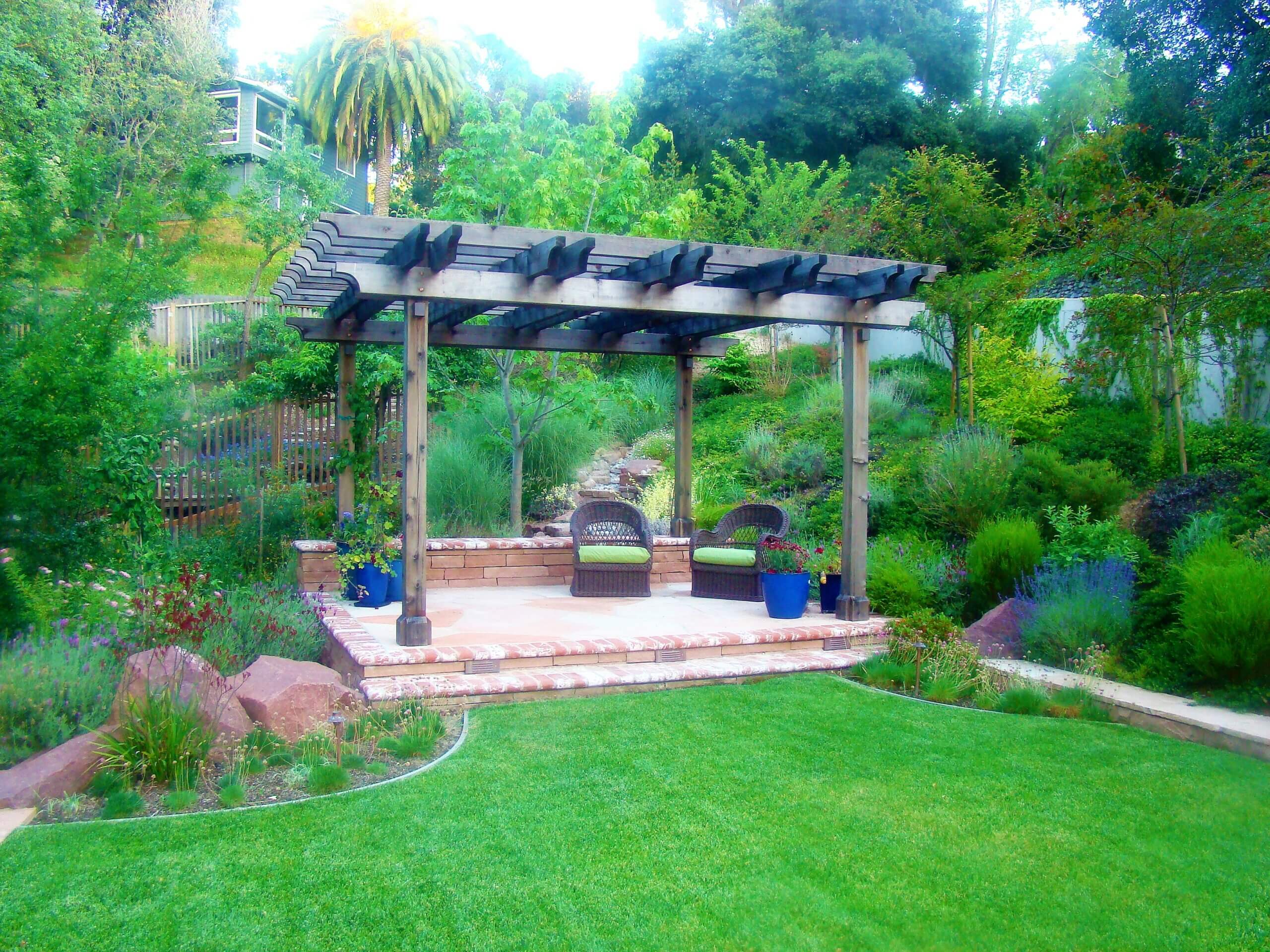 Pergola built over brick raised patio with planter pots and garden seating in a lush green California backyard environment