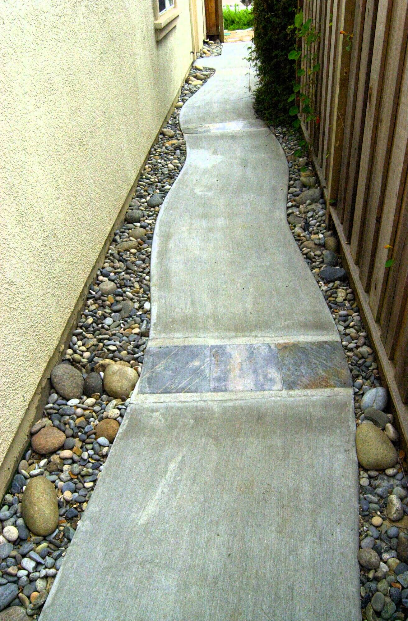 Curved and textured concrete pathway running down a narrow side yard with rocks and gravel on either side
