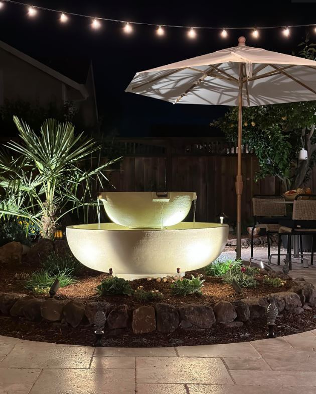 Night view of patio with custom round stone water feature with uplights surrounded by plantings